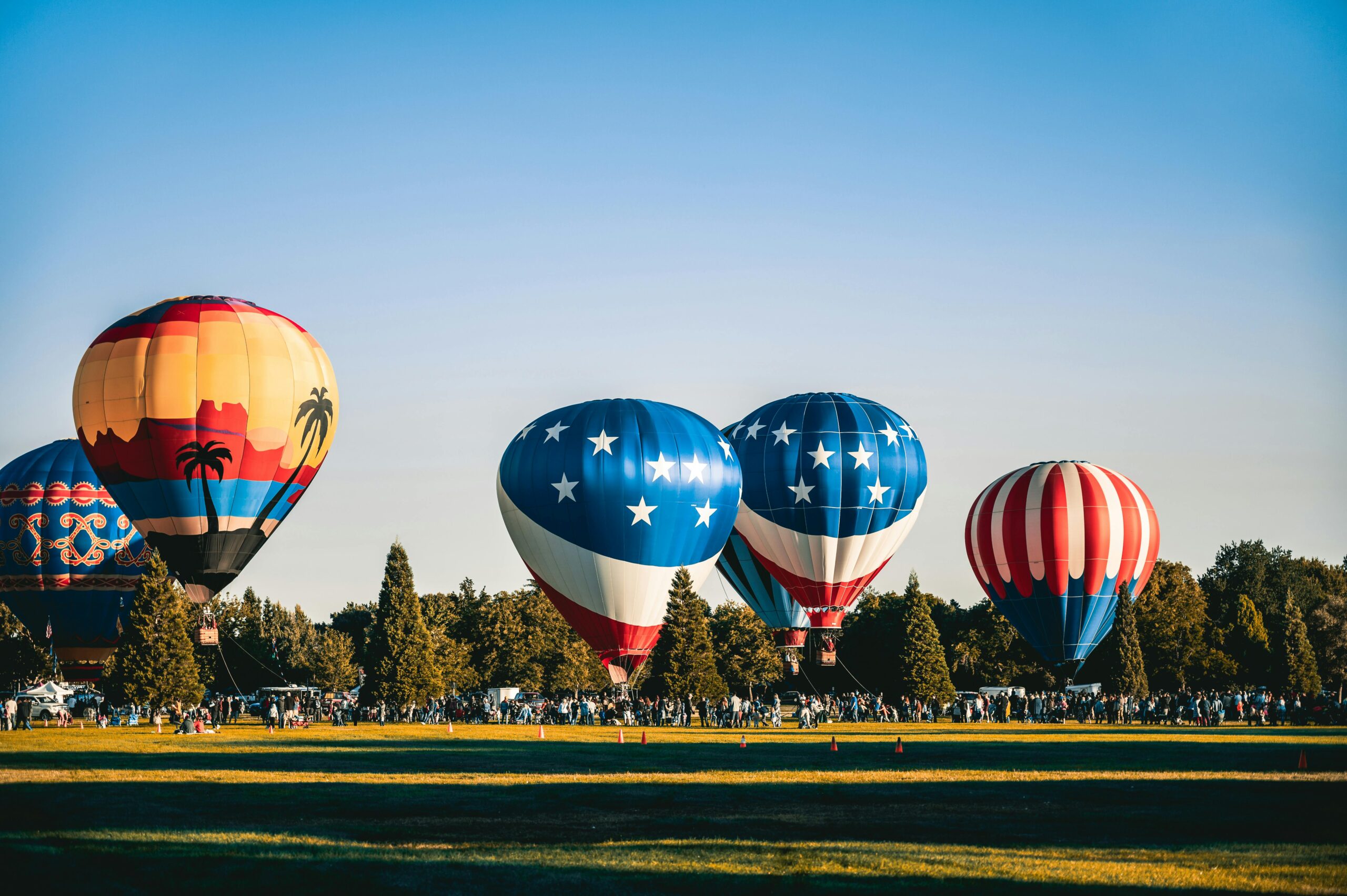 Brightly colored hot air balloons soar over a festival in an open field during a sunny day.
