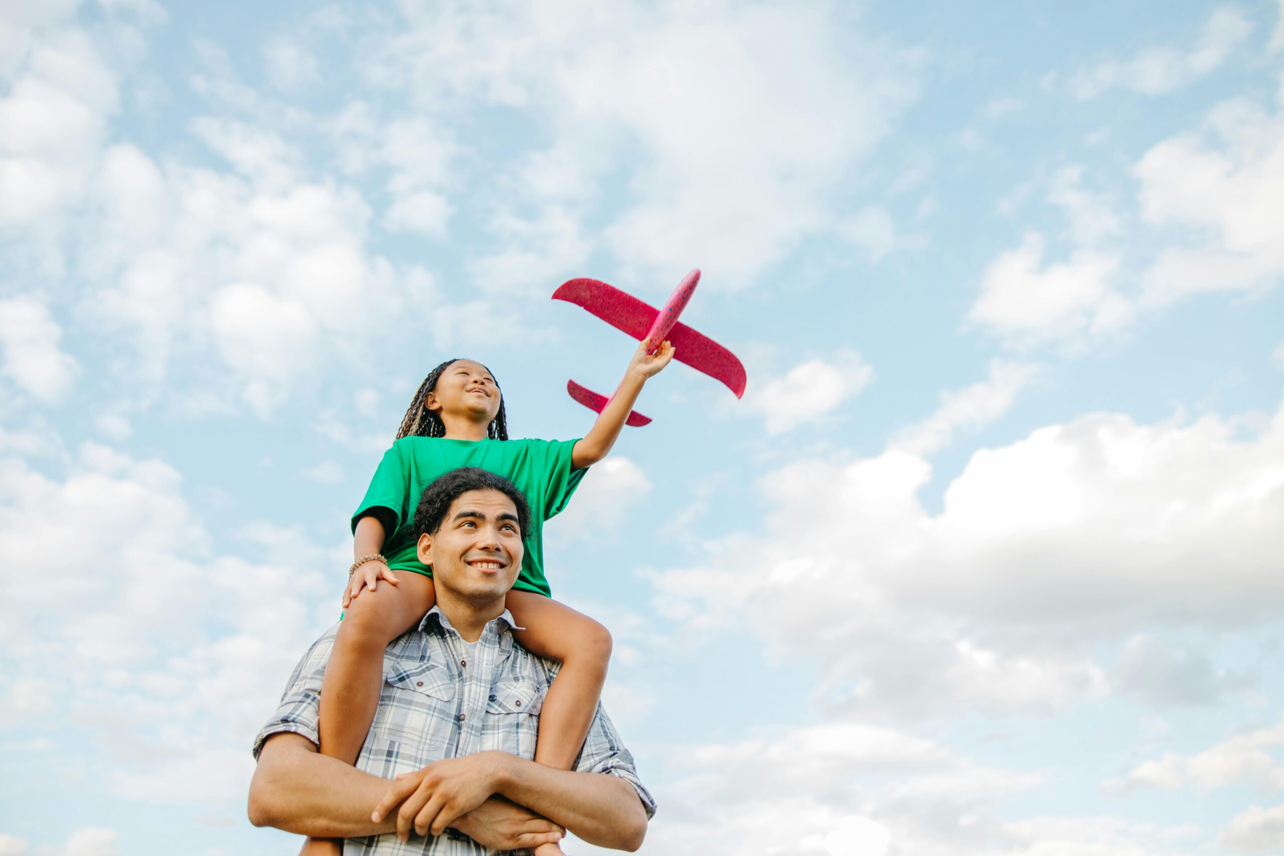 A young girl on her father's shoulders holding a toy airplane against a bright sky.